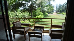 A balcony with wooden furniture and a glass table, overlooking a lush garden with palm trees and a pathway. - Laguna Beach Resort Murud (Dapoli)