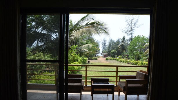 A room with a sliding glass door leading to a balcony with a view of a garden and palm trees. - Laguna Beach Resort Murud (Dapoli)