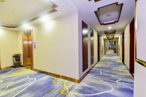 Un couloir d’hôtel avec un tapis à motifs, un ascenseur moderne et un long couloir bordé de portes des deux côtés.