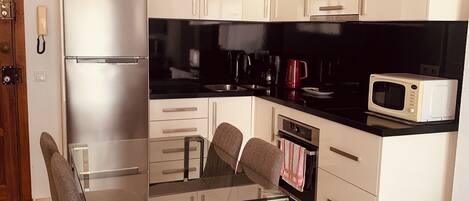 A modern kitchen with white cabinets, a stainless steel refrigerator, a black countertop, and a glass dining table with four chairs.