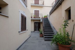 Un patio estrecho con una escalera que sube a un piso superior, plantas en macetas y una puerta en el centro.