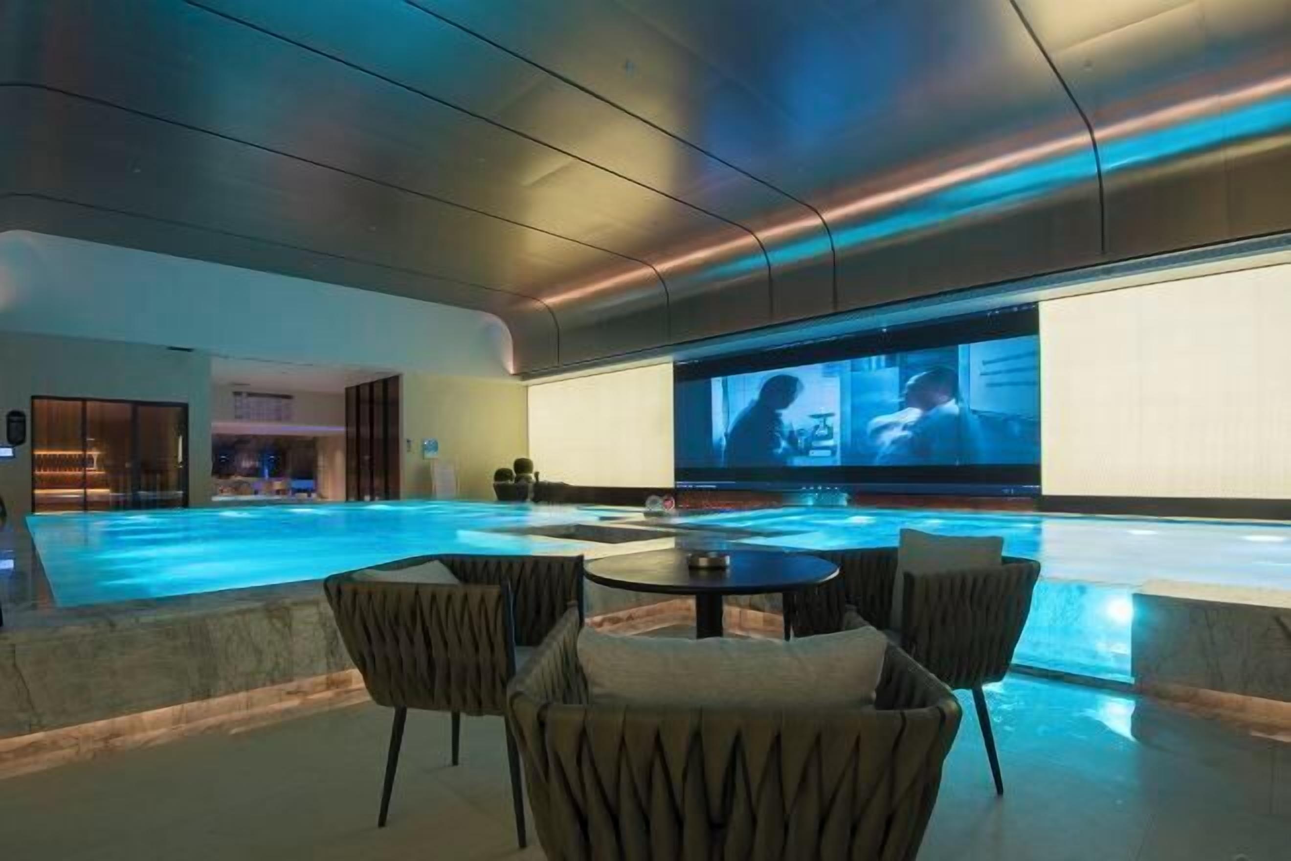 Ein moderner Poolbereich im Innenbereich mit einer großen Leinwand, die ein Sportereignis zeigt, umgeben von bequemen Sitzgelegenheiten und einem eleganten Deckenbau.