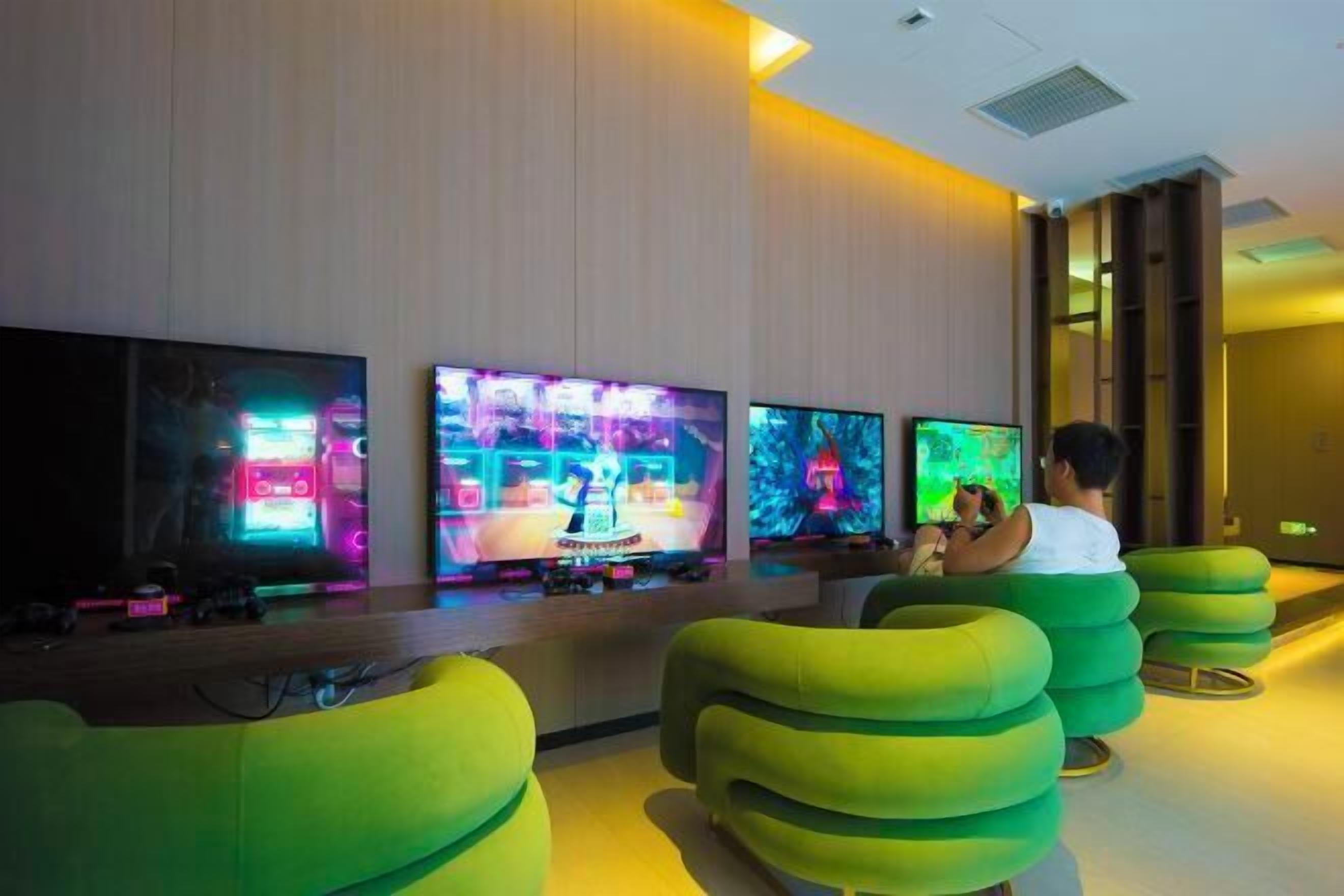 Un hall d’hôtel moderne doté de plusieurs téléviseurs à écran plat fixés au mur, de sièges confortables aux coussins verts et d’un comptoir d’accueil en bois.