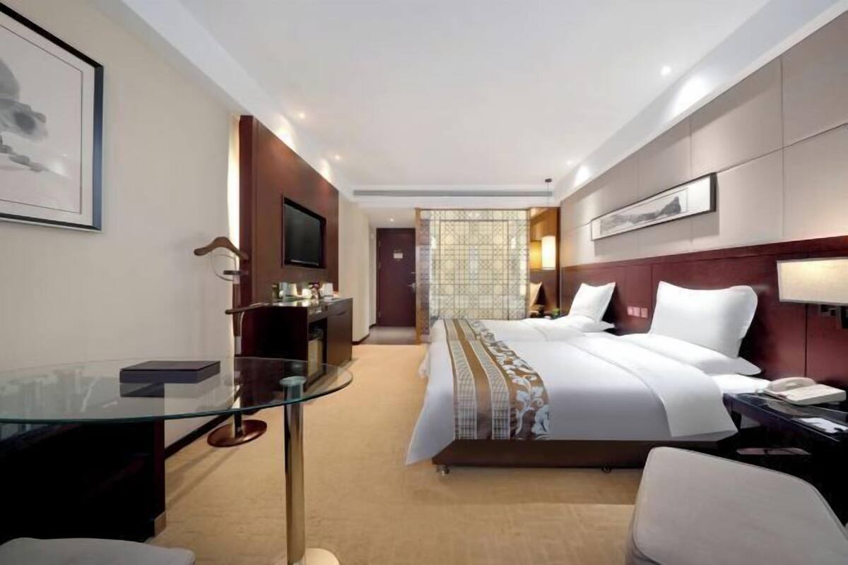 Una camera d'hotel moderna con una scrivania in vetro, un letto con lenzuola bianche, un poggiatesta in legno e un comodino con una lampada.