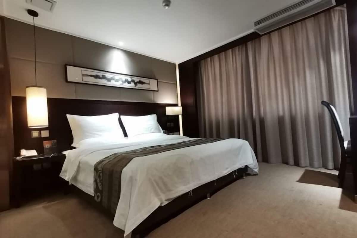 A hotel room with a large bed, a headboard with a framed picture, bedside lamps, and a large window with curtains.