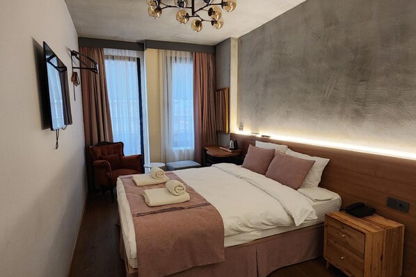 Une chambre d’hôtel avec un grand lit, une table de chevet en bois, un bureau avec une chaise, un téléviseur fixé au mur et une fenêtre avec des rideaux.