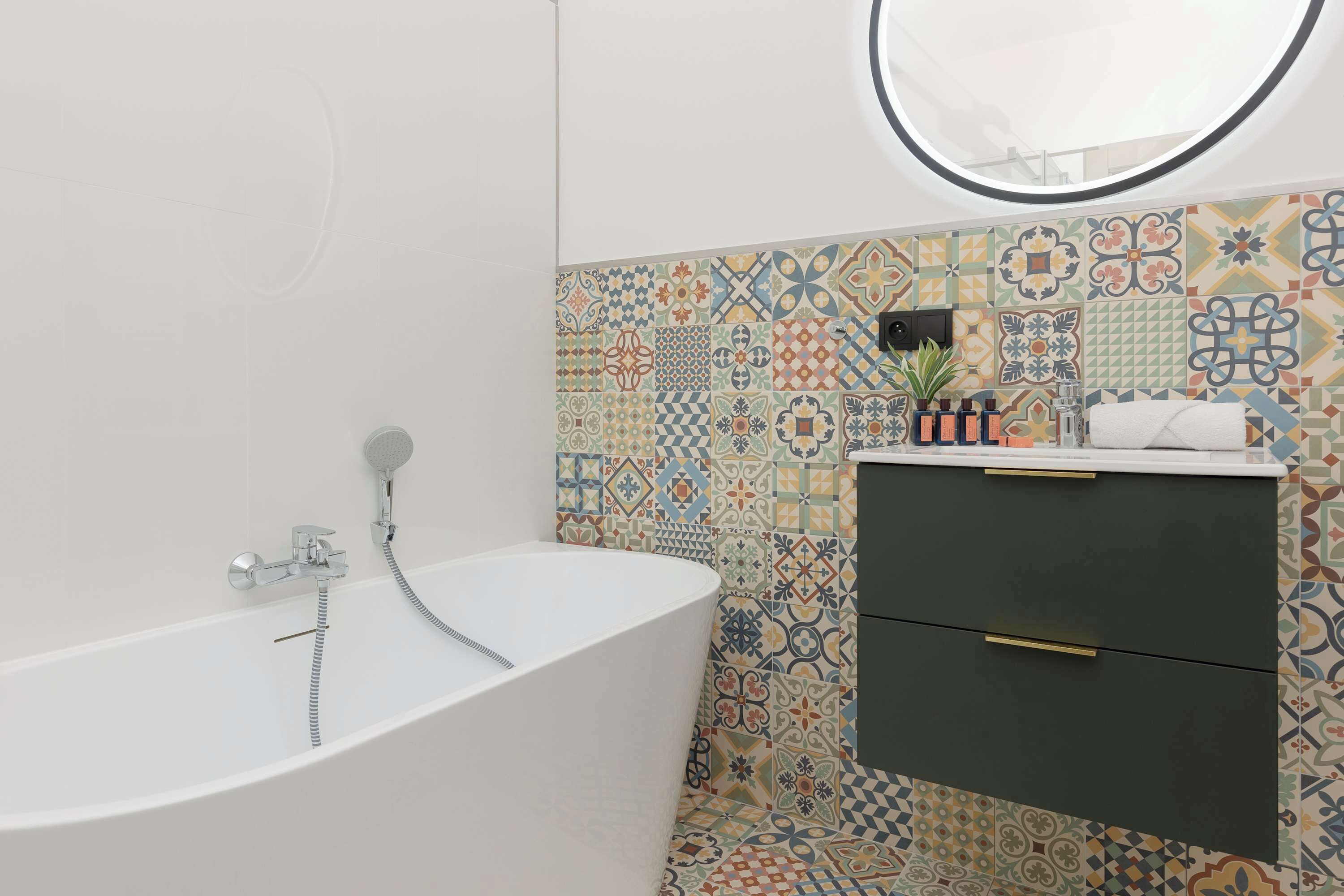 Een moderne badkamer met een vrijstaand bad, een ronde spiegel erboven en een betegelde muur met een kleurrijk patroon.