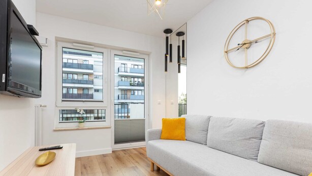 A modern living room with a grey sofa, a small wooden table, a mounted TV, and a large window with a view of a building.
