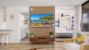 A modern living room with a wooden TV stand, a sofa, a dining area, and a kitchen. - Breezy condo w/ pool & beach access (Honolulu)
