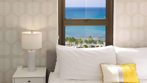 A bed with white bedding, a yellow pillow, a nightstand with a lamp, and a window with a view of the ocean and palm trees. - Breezy condo w/ pool & beach access (Honolulu)