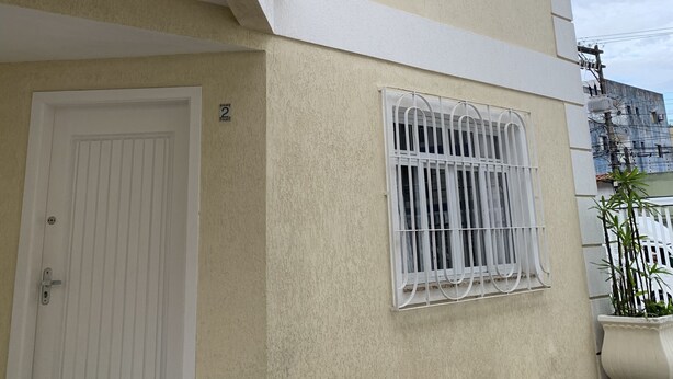 A two-story building with a beige exterior, white trim, a door, and a window with metal bars. - Cozy townhouse in gated community for families - strategic point in Enseada (Guarujá)