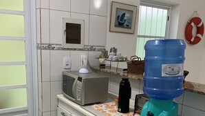 A kitchen with white cabinets, a microwave, a coffee maker, a water dispenser, and a window with blinds. - Cozy townhouse in gated community for families - strategic point in Enseada (Guarujá)