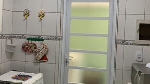 A kitchen with white tiled walls, a white stove, a microwave, and a door with frosted glass panels. - Cozy townhouse in gated community for families - strategic point in Enseada (Guarujá)