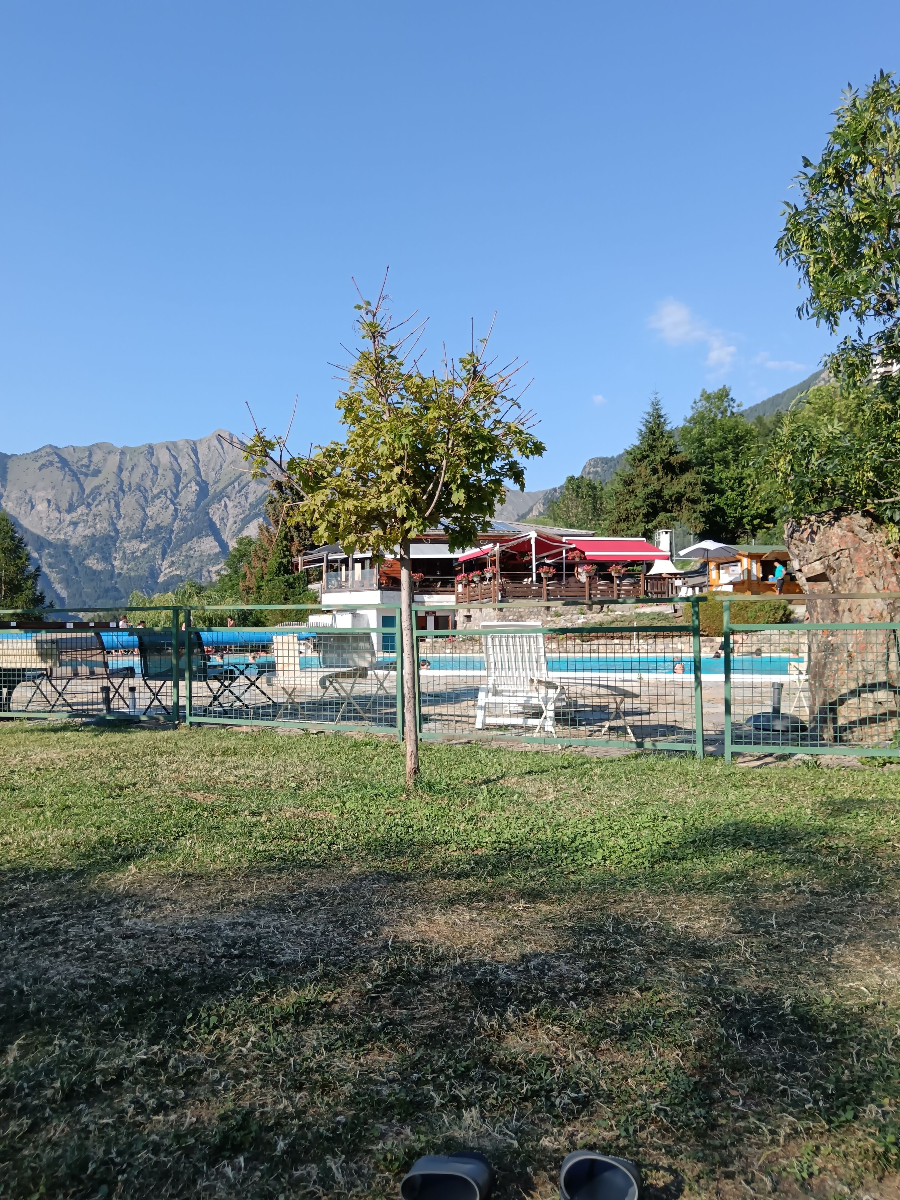 Un espace piscine entouré d’une clôture, un bâtiment avec un toit rouge et des montagnes en arrière-plan.