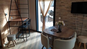 Ein modernes Zimmer mit Holzboden, einem runden Tisch mit Stühlen, einem kleinen Hocker, einer Leiter, einem an der Wand befestigten Fernseher und einem Fenster mit Vorhängen.
