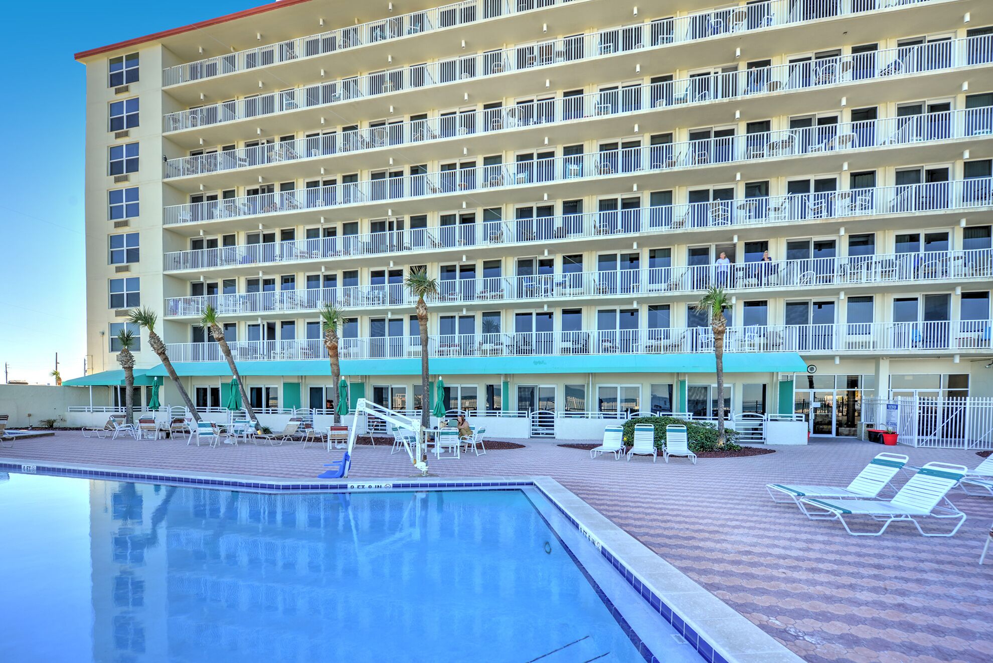 Een meerlaags hotel met een zwembad, terras en palmbomen.
