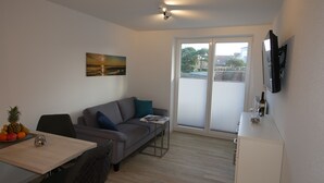 Un salon moderne avec un canapé gris, une petite table sur laquelle se trouve un ordinateur portable, un téléviseur fixé au mur et une porte-fenêtre coulissante donnant sur un espace extérieur.