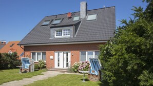 Een twee verdiepingen tellend huis met een schuin dak, een schoorsteen en een kleine tuin met gazon en twee blauw gestreepte stoelen.