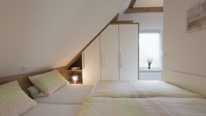 Een slaapkamer met een schuine kap, houten balken, een raam met zonwering en een nachtkastje met een lamp.
