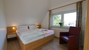 A bedroom with a bed, bedside lamps, a window with curtains, a chair, and a vase with flowers. - APARTMENT 2 ON THE 2ND FLOOR (Norderney)