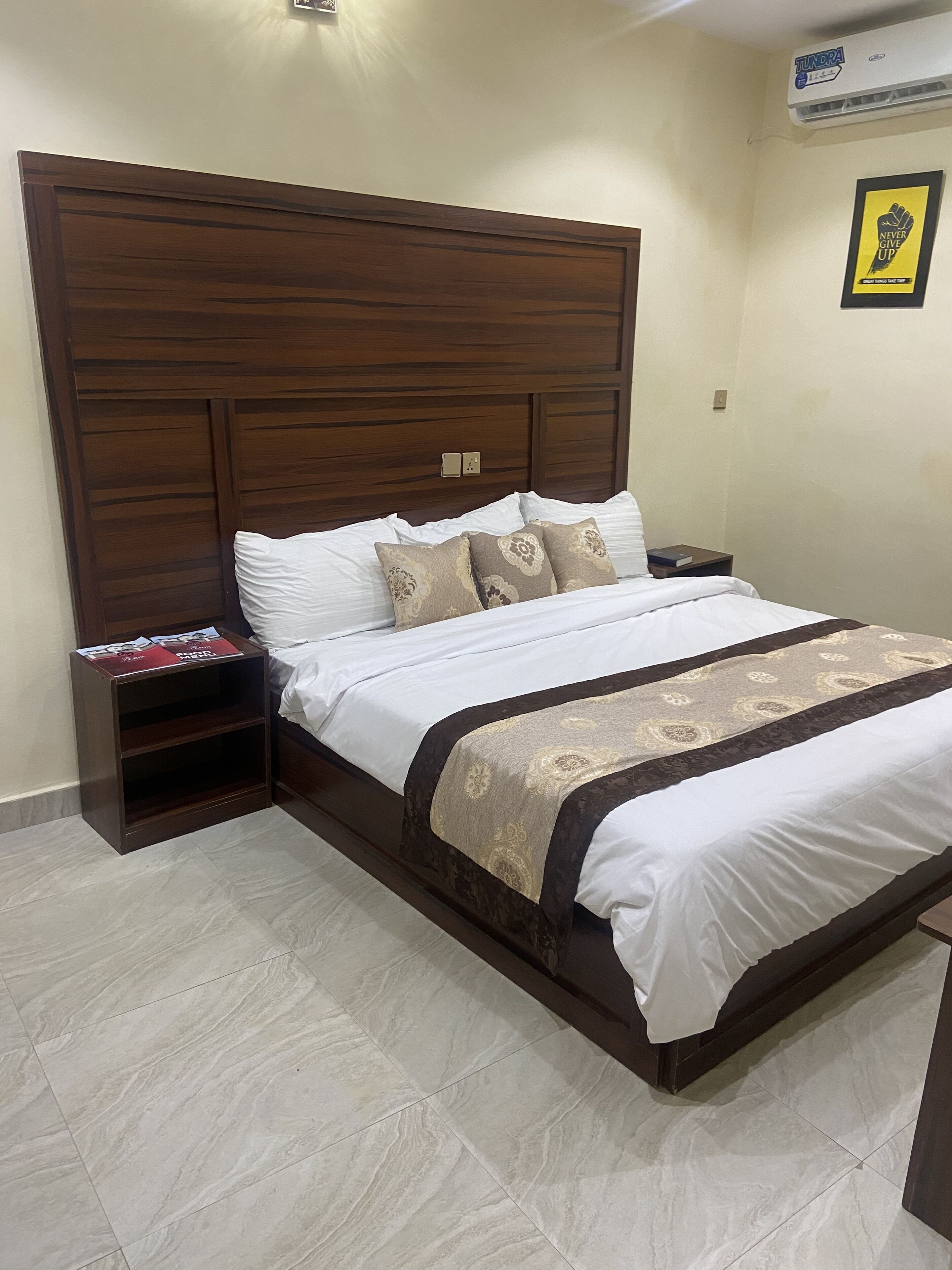 Una cama bien hecha con cabecera de madera, una mesita de noche con un libro y un aire acondicionado de pared.
