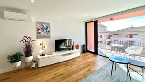 A modern living room with a flat-screen TV, white console, and potted plants. A balcony with outdoor furniture is visible. - One - Luxurious apartment by HD PROPERTIES (Vilamoura)