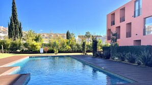 A swimming pool with a wooden deck, surrounded by greenery and a pink building. - One - Luxurious apartment by HD PROPERTIES (Vilamoura)