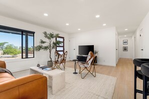 A modern living room with a large window, a flat-screen TV, and a seating area with a sofa and two chairs.
