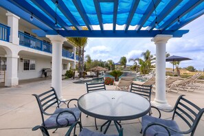 Une terrasse extérieure avec une table ronde en verre et des chaises, une piscine, des chaises longues et une pergola bleue.