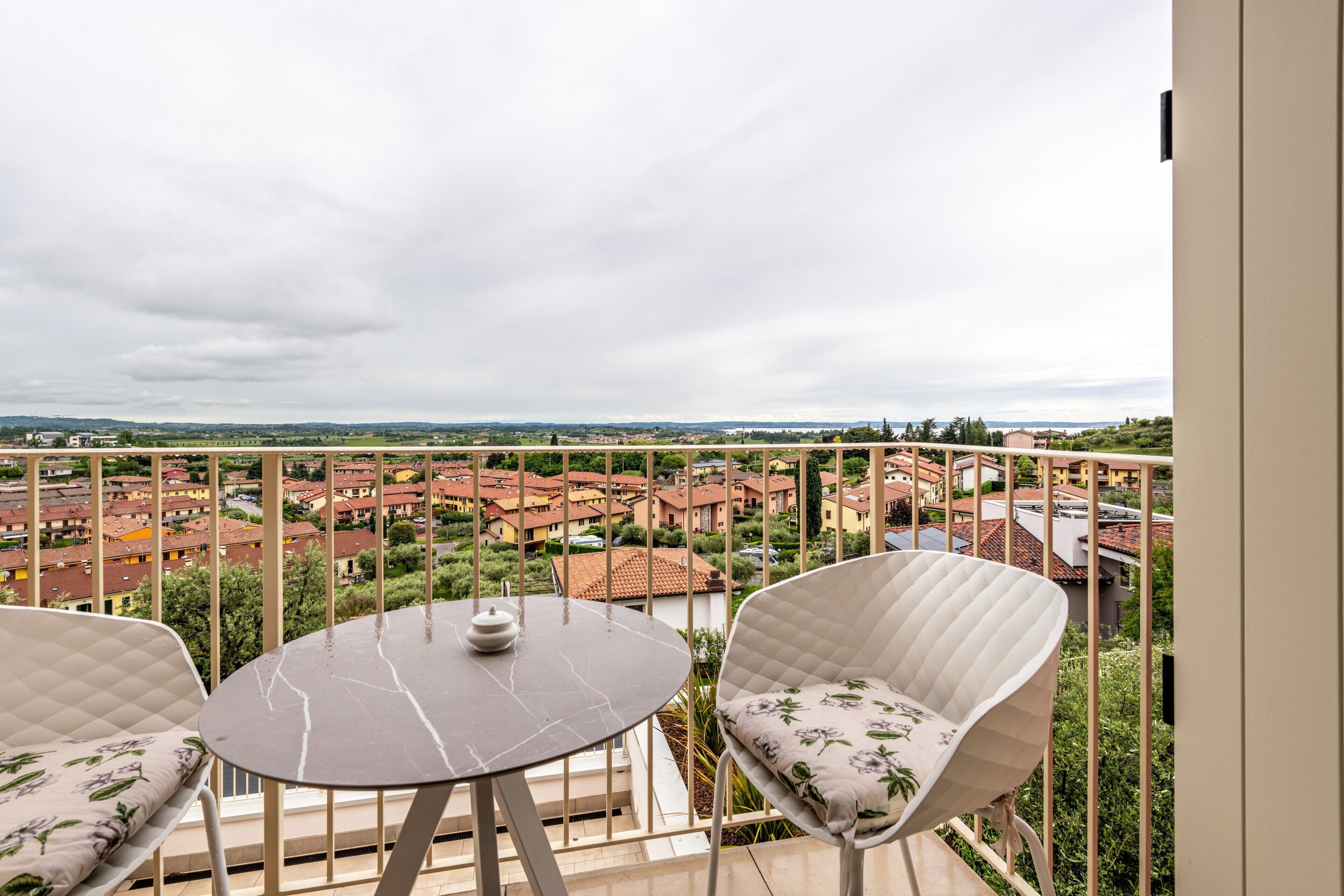 A balcony with a round table and two chairs, overlooking a residential area with red-roofed buildings.