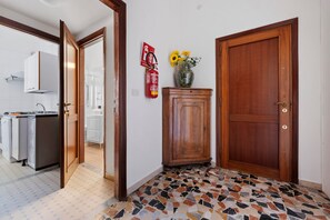 A hallway with a tiled floor, a wooden door, a cabinet with a vase of flowers, and a fire extinguisher on the wall.