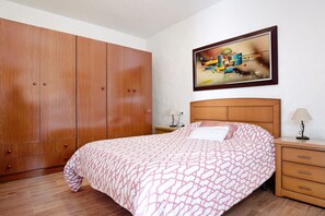 A bedroom with a large wooden wardrobe, a bed with patterned bedding, a nightstand with a lamp, and a framed abstract painting on the wall. - Vacation Home "Las Niñas" with Sea View, Private Terrace & Garden (Vejer de la Frontera)