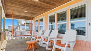 Ein überdachter Außenbereich mit weißen Stühlen und orangefarbenen Kissen, einer Holzterrasse und großen Fenstern.