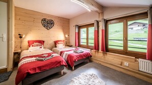A wooden cabin bedroom with two beds, a heart-shaped wall decoration, and a view of a green landscape through a large window. - Apartment 'Coeur De Charmy' with Mountain Views and Wellness Spa (Abondance)