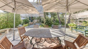 En patio med et rundt bord og stole, omgivet af store parasoller, med grøn vegetation og en stenvæg i baggrunden.