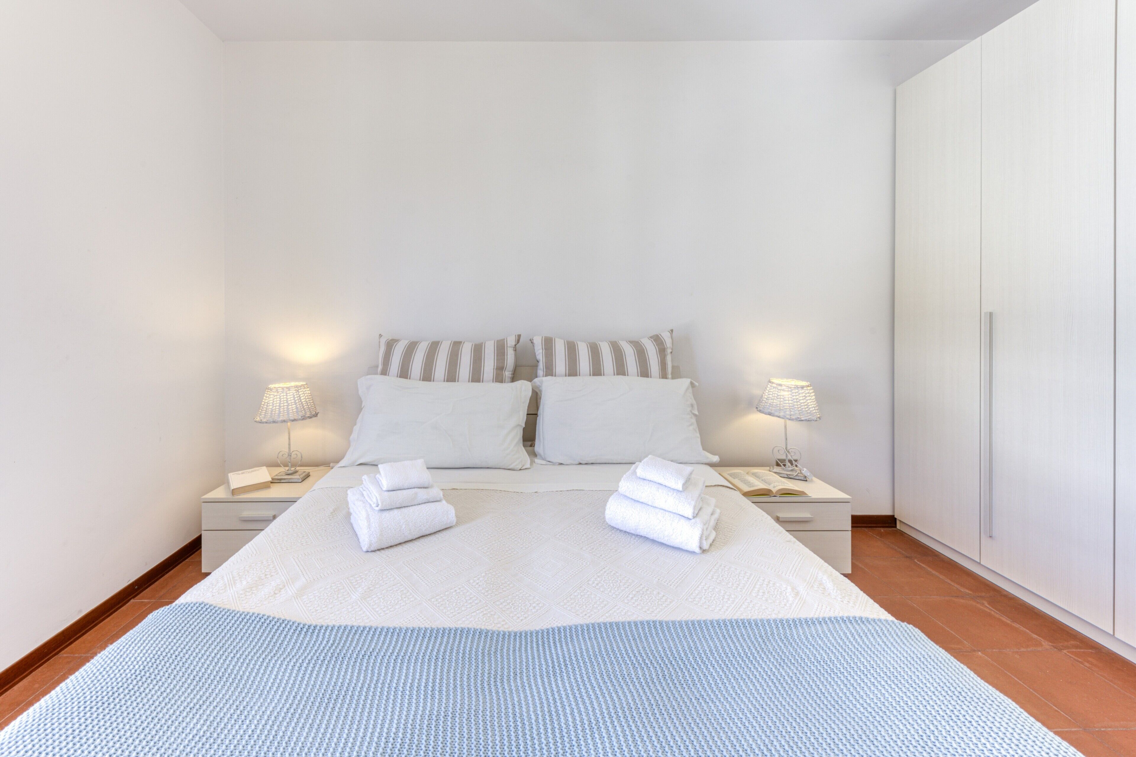 A neatly made bed with pillows and folded towels, a bedside table with a lamp, and a wardrobe.