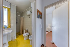 Une salle de bain avec un sol jaune, un lavabo, deux toilettes, une douche et une porte donnant sur une autre pièce.