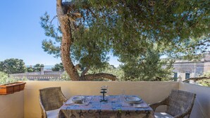 An outdoor dining area with wicker chairs, a table set with plates, glasses, and a pitcher, under a large tree with a view of buildings in the background.