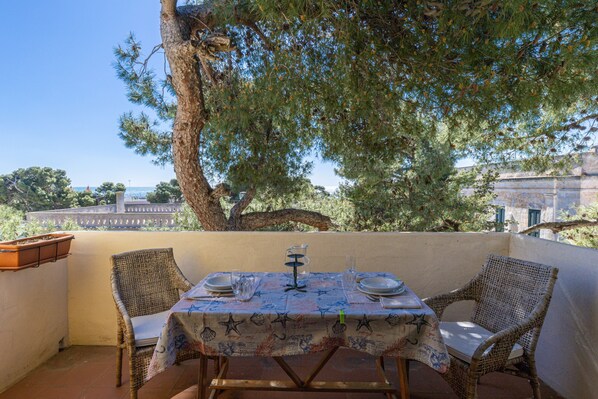Un espace de restauration en plein air, avec des chaises en osier, une table dressée avec des assiettes, des verres et une carafe, situé sous un grand arbre, d’où l’on peut admirer des bâtiments en arrière-plan.