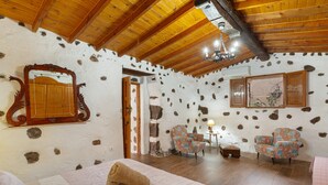 A rustic room with a stone wall, wooden ceiling beams, a bed with a pink quilt, two patterned armchairs, and a small table with a lamp.