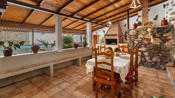 An outdoor dining area with a stone fireplace, wooden ceiling, and potted plants.