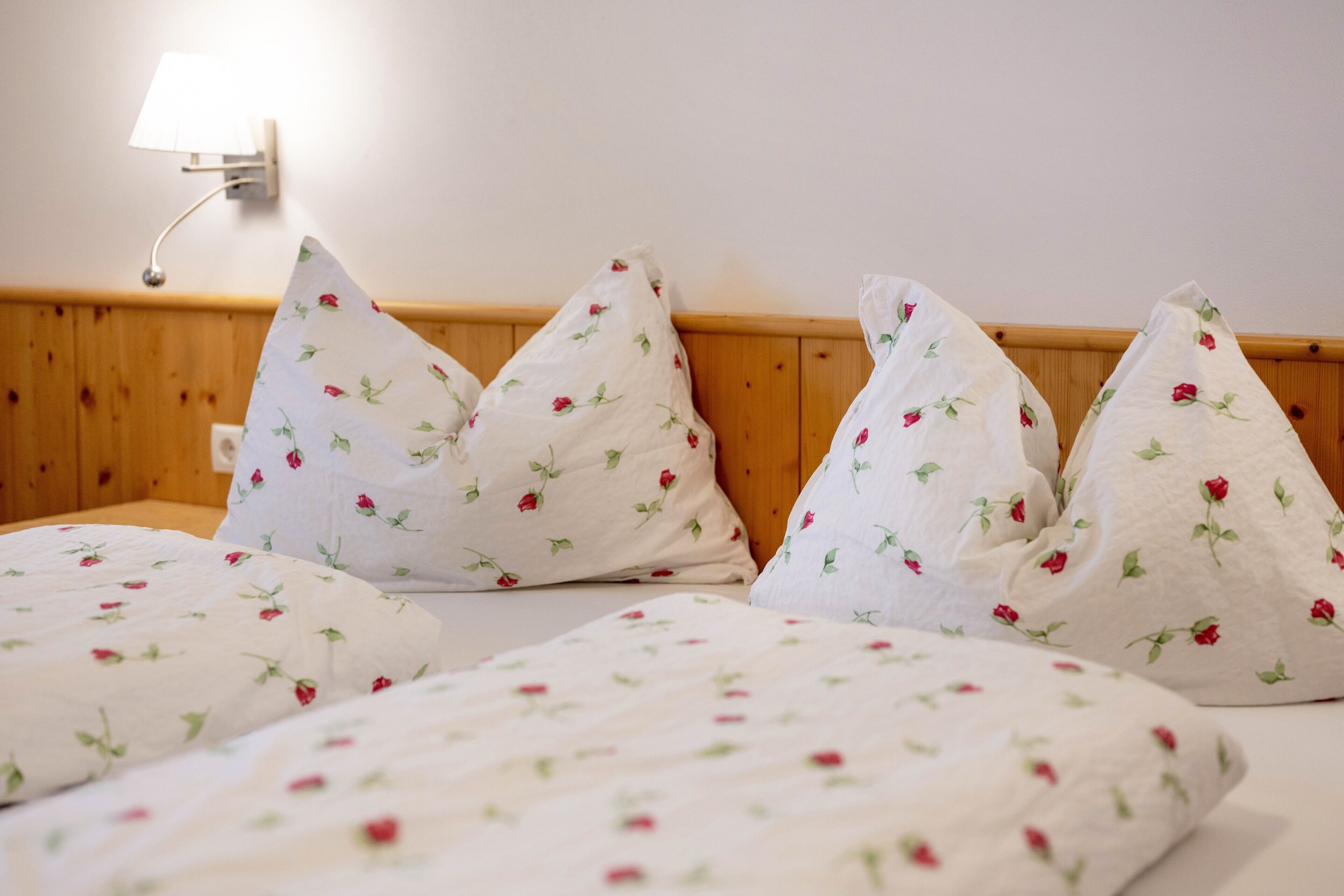 A bed with floral-patterned bedding and pillows, a wooden headboard, and a wall-mounted lamp.