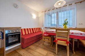 A kitchenette with an oven, a red sofa, a wooden dining table with a red tablecloth, and a window with sheer curtains.