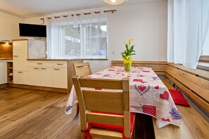 Un rincón comedor con una mesa dispuesta para dos personas, una silla de madera, un florero con flores y una ventana con cortinas.