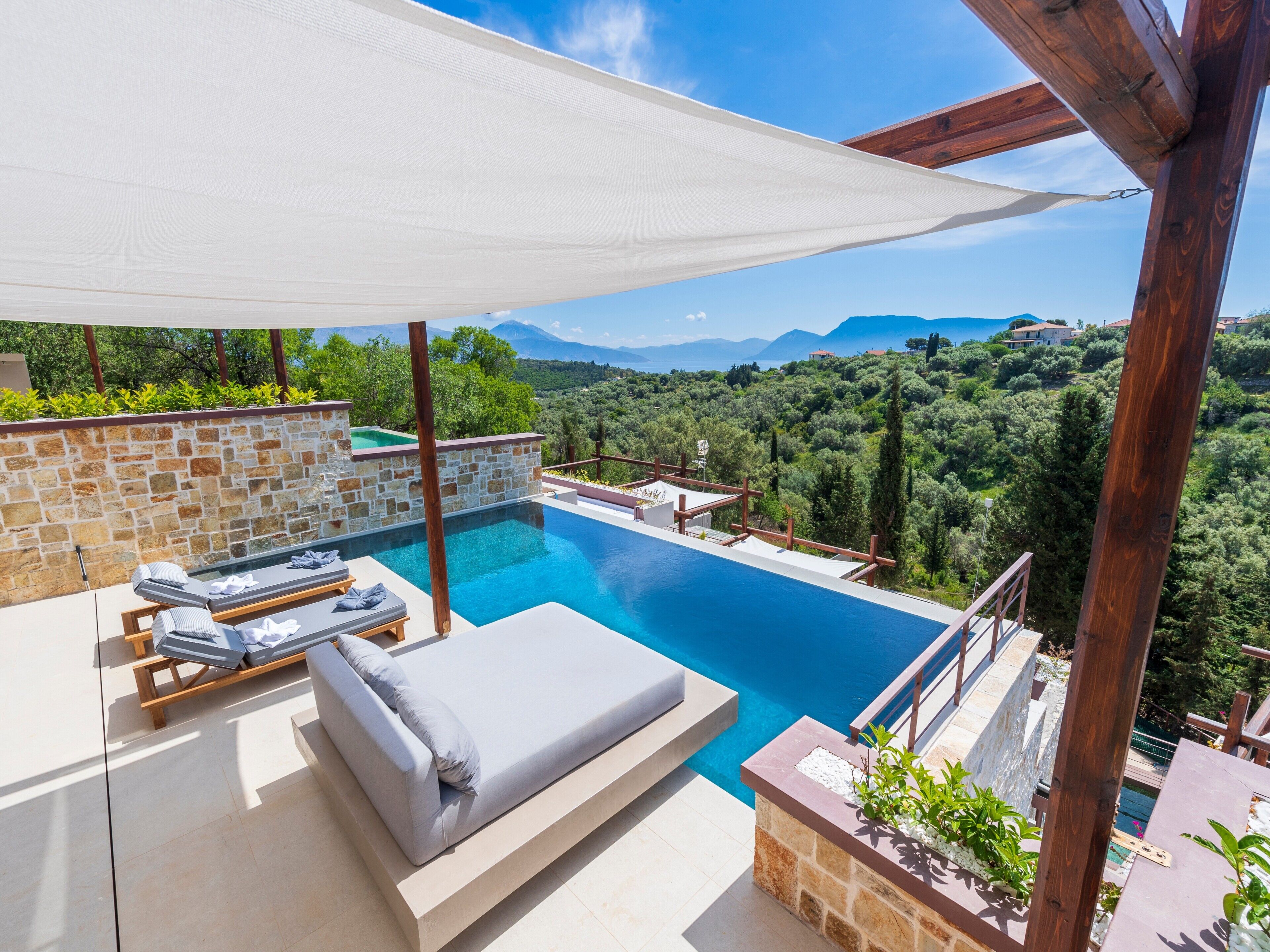 Una piscina con muro de piedra y cubierta de madera, un área de descanso con cojines, y vista a un paisaje montañoso.