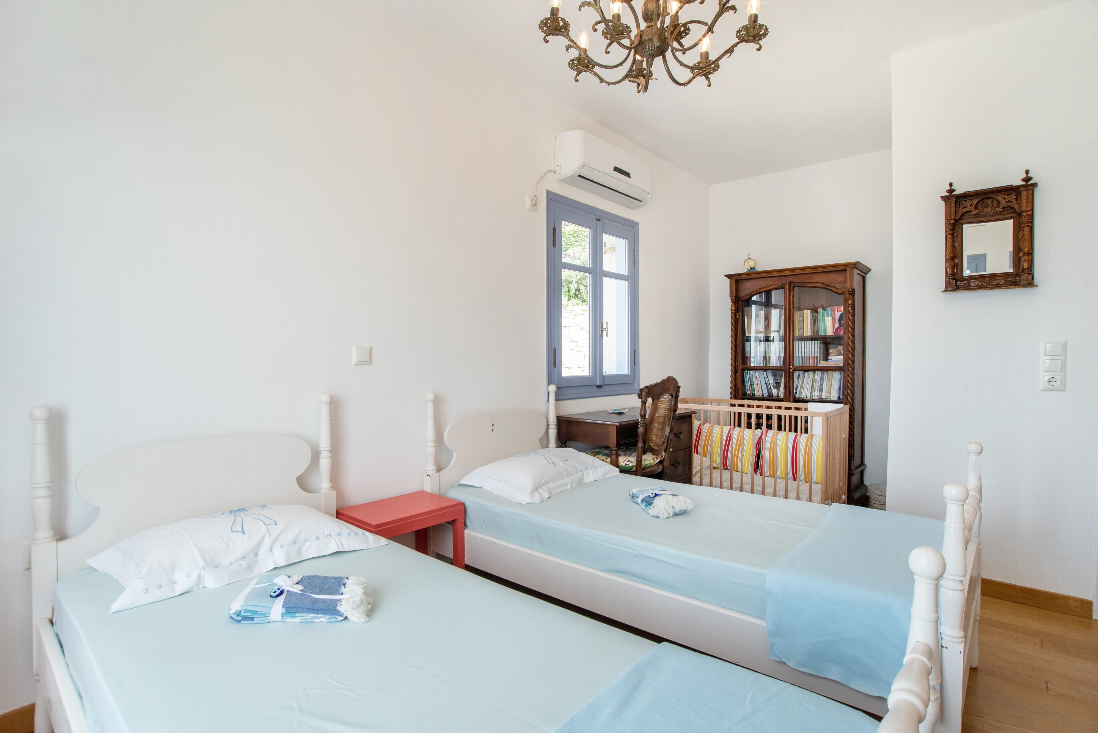 Une chambre avec deux lits, une table de chevet rouge, une armoire en bois remplie de livres et une fenêtre avec un appareil de climatisation.