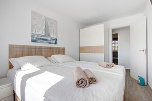 A neatly made bed with rolled towels, a wooden headboard, a painting of a sailboat, and a wardrobe with a wooden accent.
