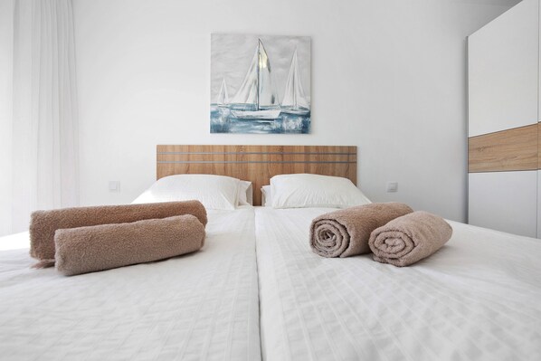 Habitación de hotel con cama, dos toallas enrolladas, cabecera y un cuadro de un velero en la pared.