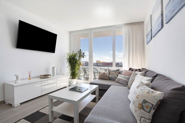 A modern living room with a flat-screen TV, a white coffee table, a grey sofa, and a large window with a city view.