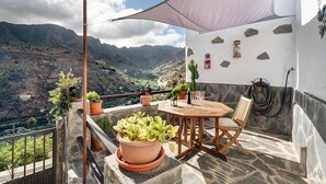 Una terraza con mesa y sillas, plantas en macetas, pared blanca con detalles de piedra y vistas a las montañas.